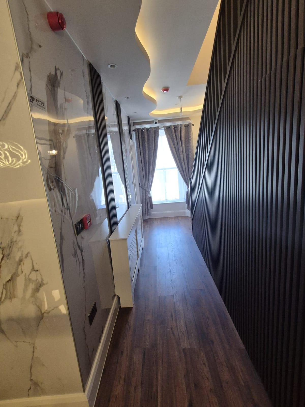 A hallway with a white marble-effect wall, dark slatted panelling, a curved glowing ceiling and a curtained window