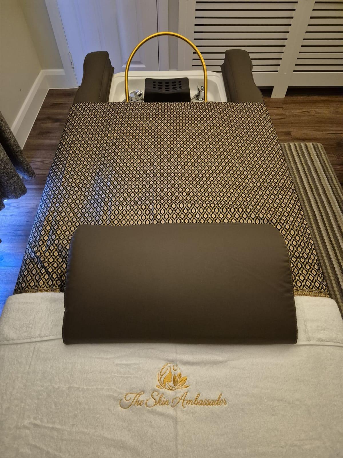 A head spa bed with gold-patterned silk, a dark bolster and a white towel embroidered with The Skin Ambassador logo
