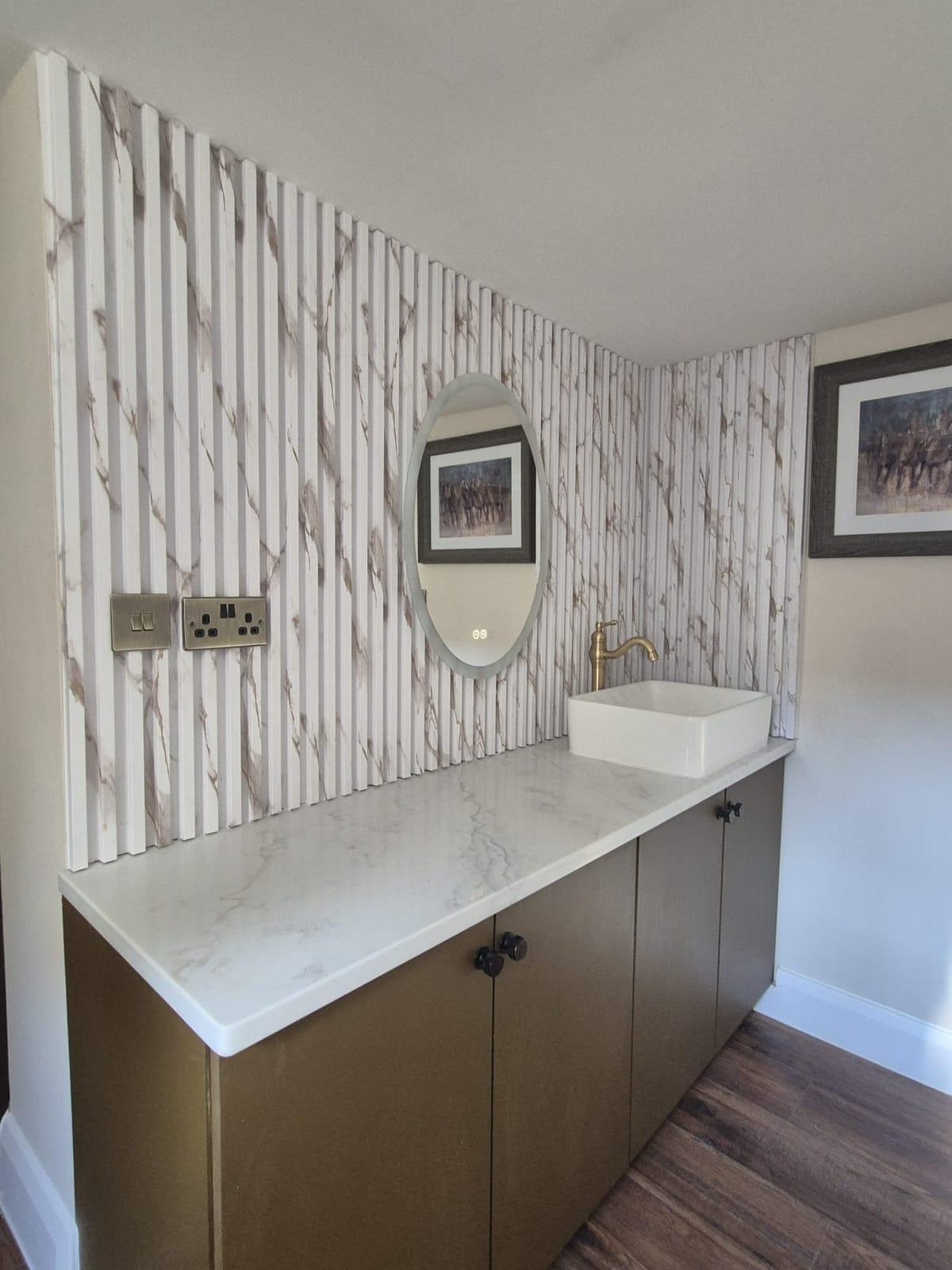 A white marble-effect fluted wall with an oval mirror, a vessel basin and brass tap on a marble counter