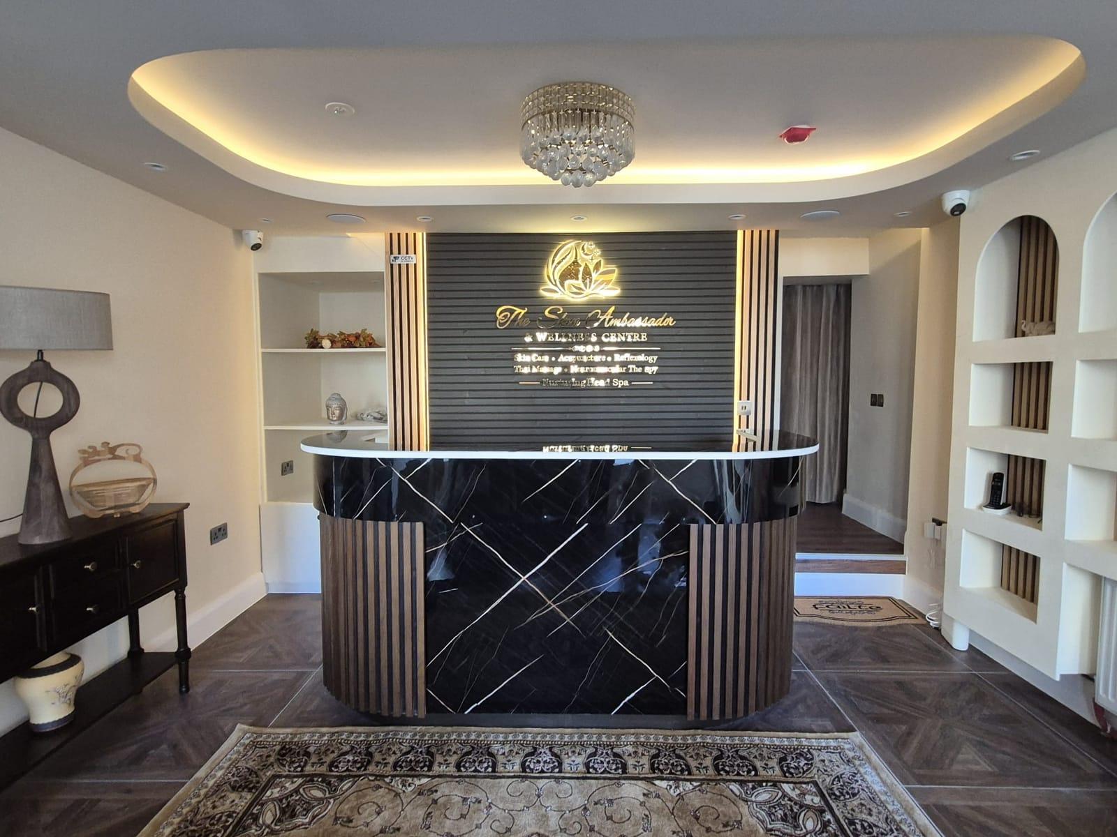 The reception desk in black and gold marble with wooden slats, beneath a crystal chandelier, in front of the illuminated Skin Ambassador sign