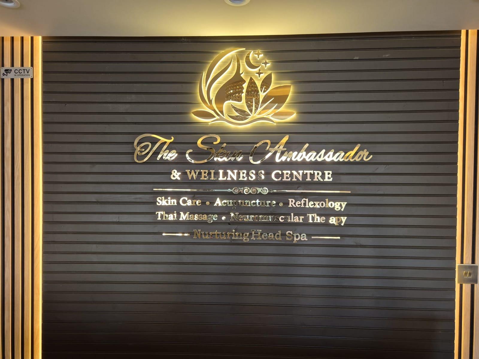 The illuminated gold Skin Ambassador & Wellness Centre sign listing its treatments on a dark slatted wall