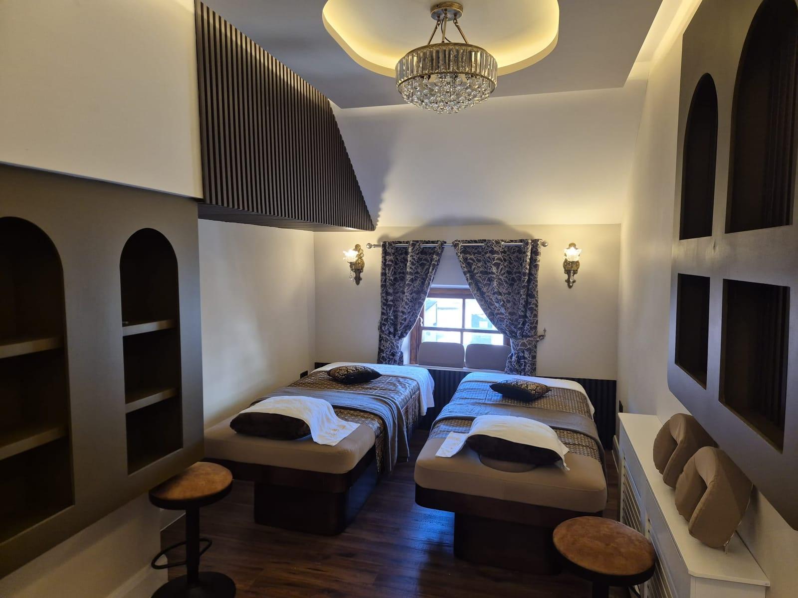 Two treatment beds with silk throws and cushions under a crystal chandelier, between arched alcoves and brass wall lights
