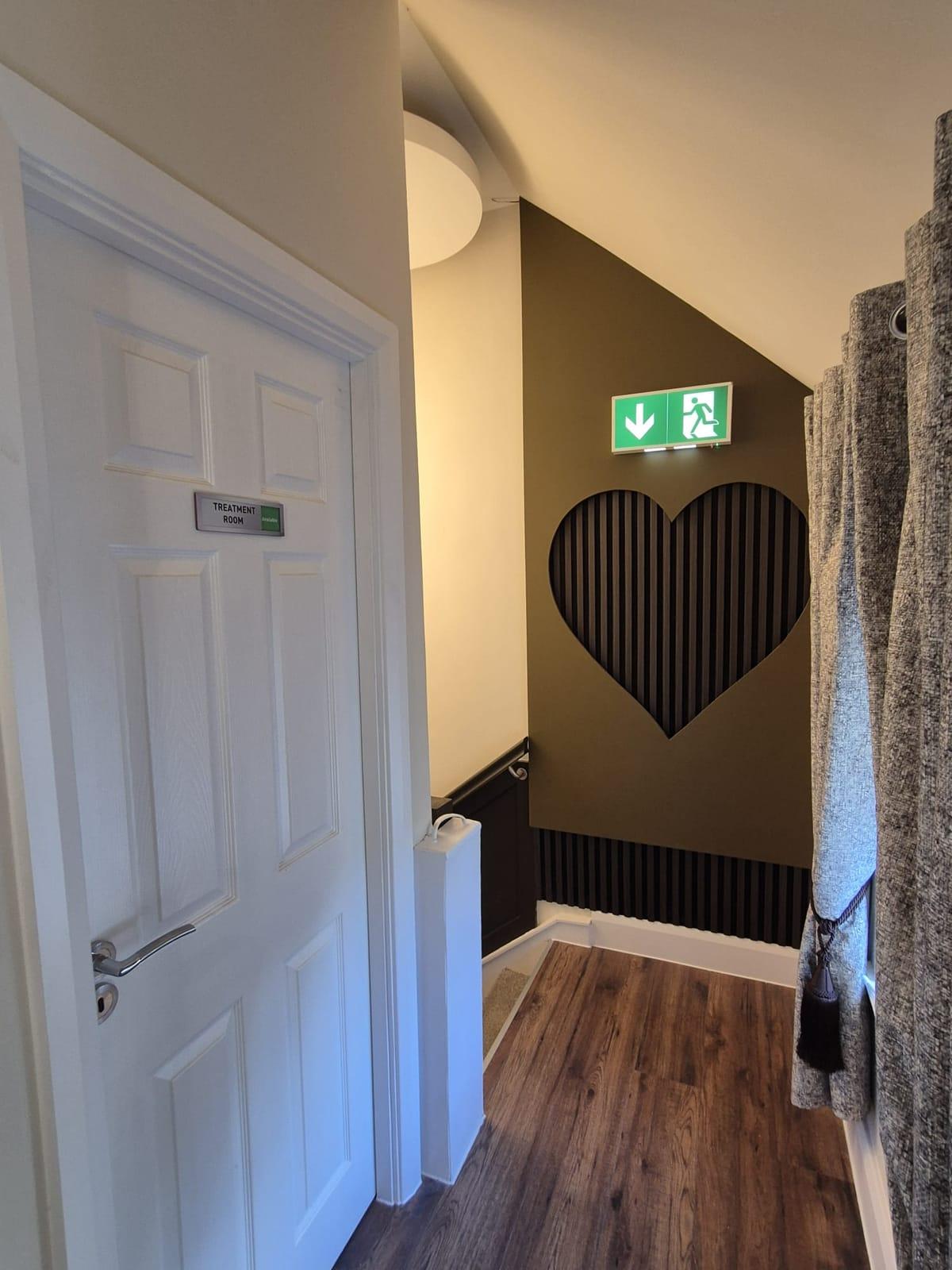 An upstairs landing with a heart-shaped slatted wall feature, a treatment room door and wooden flooring