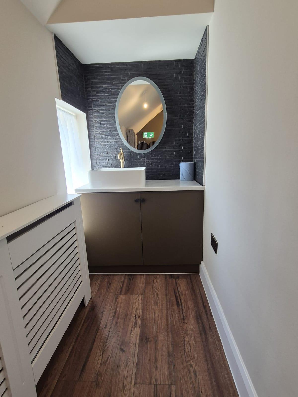 A white vessel basin with a brass tap and oval mirror against a dark slate wall